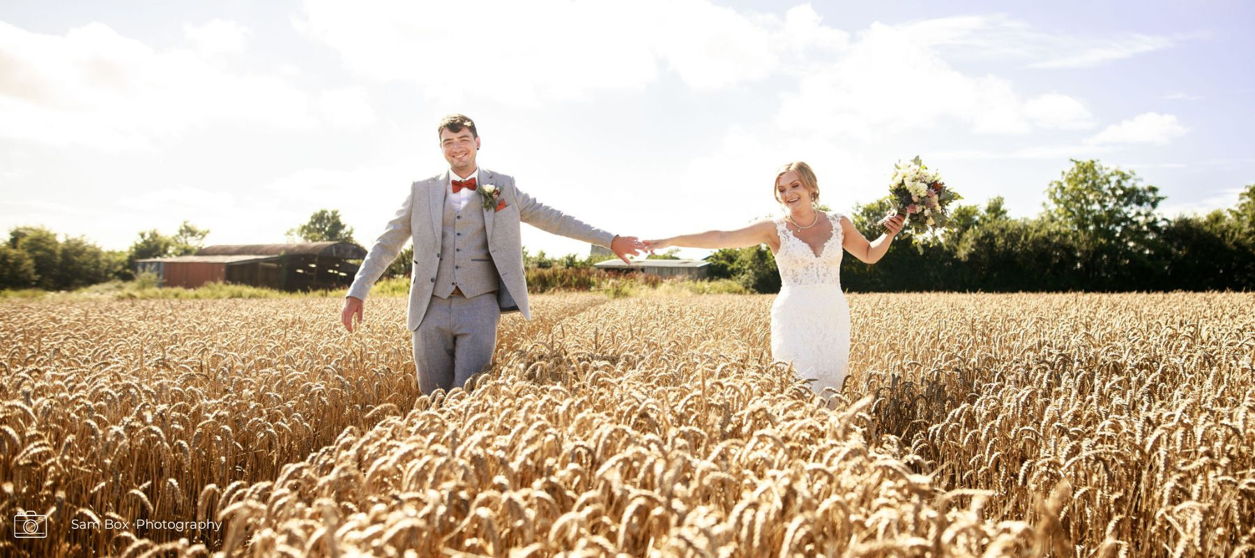 Field of wheat with bride and groom walking through with bride holding a summer bridal bouquet